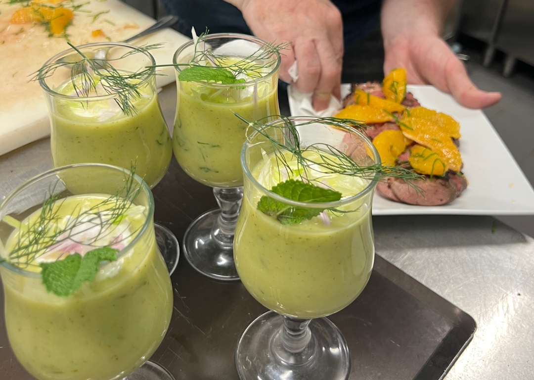 Formation en cuisine professionnelle - gros plan sur des verres de soupe verte garnis de menthe et d'aneth, préparés lors de la session, avec une assiette de viande et légumes grillés