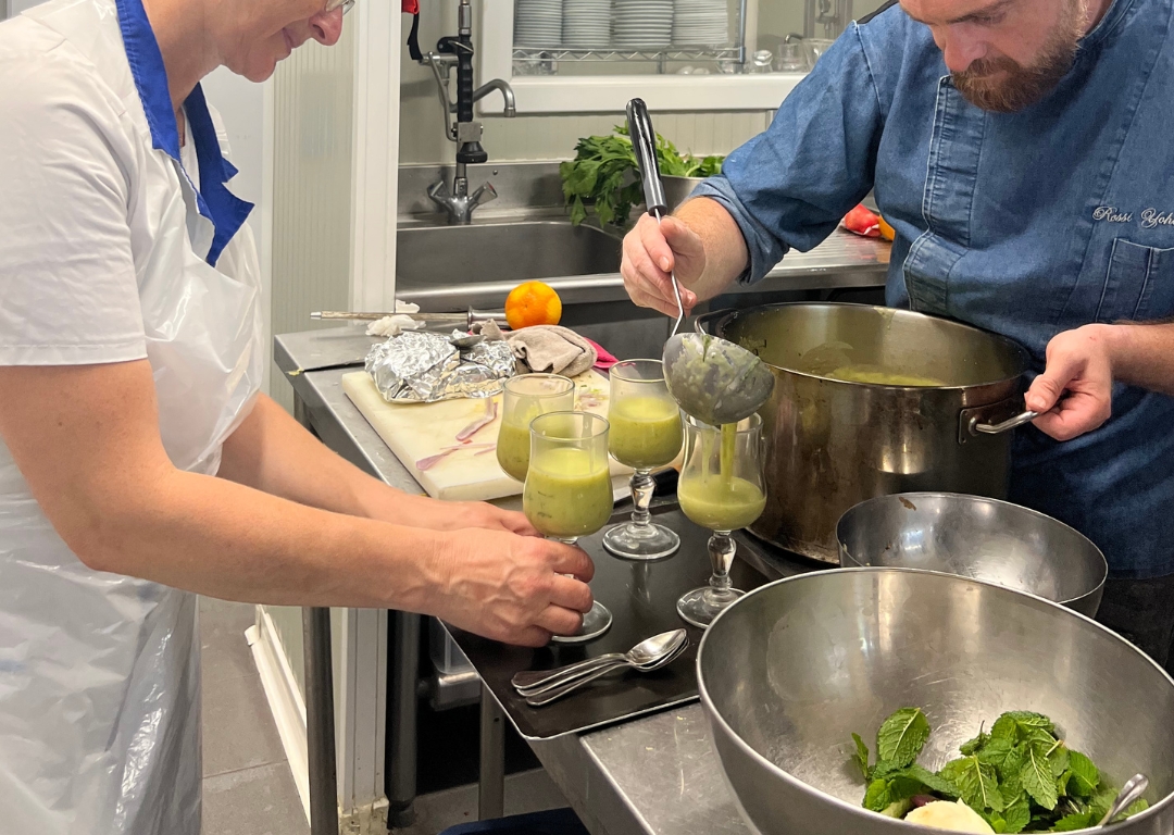 Un chef en veste bleue sert une soupe verte dans des verres avec l'aide d'une formatrice en tablier blanc.